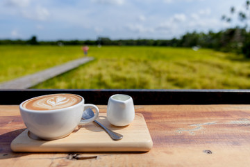  Coffee cup on wooden table and the rice field background. Cup of hot coffee with milk art on the surface placed on the wooden table.