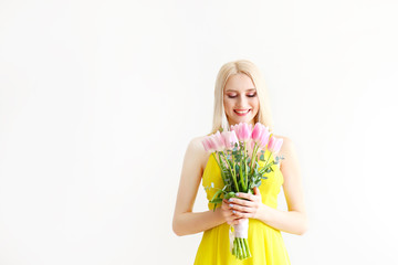 Studio portrait of gorgeous young blonde woman with long straight hair wearing bright yellow dress and holding bouquet of many tender pink tulips. Gray isolated background, copy space, close up.