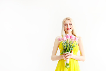 Studio portrait of gorgeous young blonde woman with long straight hair wearing bright yellow dress and holding bouquet of many tender pink tulips. Gray isolated background, copy space, close up.