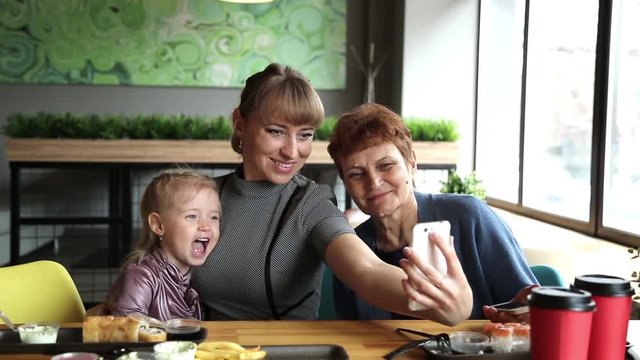Grandmother's daughter and granddaughter are photographed on the phone in a cafe. A young woman takes a picture of herself on a smartphone with an elderly mother and little daughter