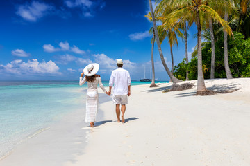 Elegant traveler couple walks down a tropical beach with coconut palm trees and turquoise waters in...
