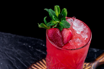 Beautiful red berry cocktail with ice, decorated with fresh berries and mint, on a round carved wooden board. Berry cocktail isolated on a black background. Close-up. Space
