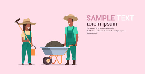 gardener man with wheelbarrow of earth woman holding hoe and bucket african american couple farmers planting flowers and vegetables gardening concept full length copy space horizontal