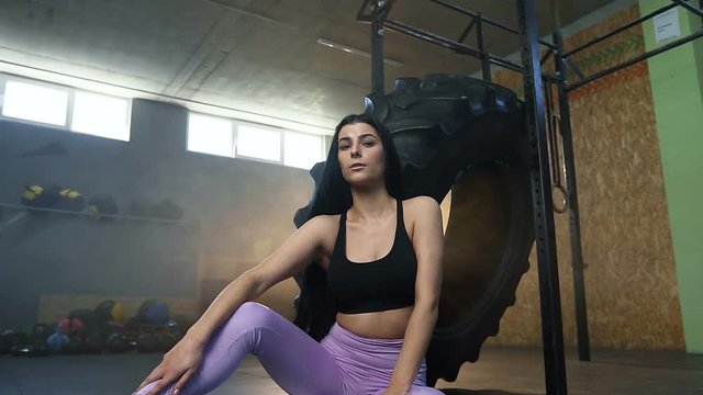 Dolly shot of attractive fit woman looking to the camera while sitting near tractor tire in the gym.