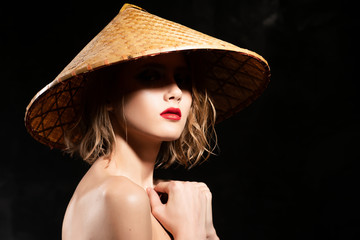 Close-up face of a beautiful young blonde girl with red lips and eyes hidden in the shadow of a cone-shaped Asian cane hat. Clean, healthy skin. Fashionable and advertising design