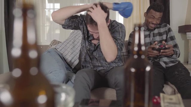 Multicultural friends watching television sitting on the couch. On the table are empty beer bottles. The guys are fooling around. Bearded young man wearing a blue boxing glove and jokingly boxing with