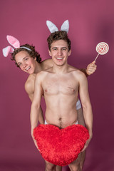 Two funny naked guys wearing bunny ears holding lollipop and big red heart over pink background