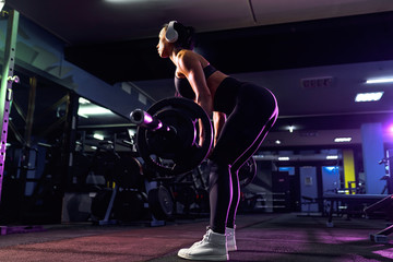 Fototapeta na wymiar Attractive fit sexy woman in wireless headphones listen music and doing crouches with a barbell in the gym. Woman training back.