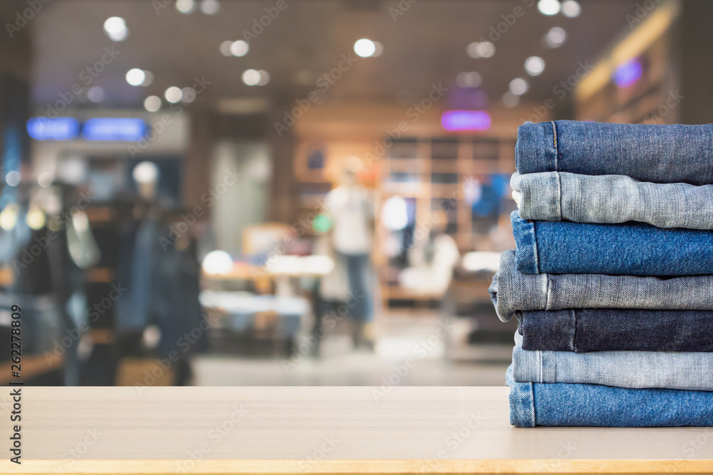 Wall mural Blue denim jeans stack on wood table top with abstract blur clothing store in modern shopping mall defocused bokeh light background