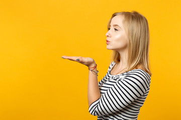 Side view of attractive young woman in striped clothes blowing sending air kiss isolated on yellow orange wall background in studio. People sincere emotions, lifestyle concept. Mock up copy space.