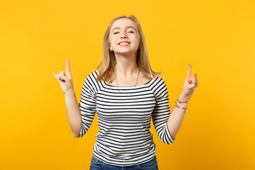 Smiling young woman keeping eyes closed, showing horns up gesture, depicting heavy metal rock sign isolated on yellow orange background. People sincere emotions, lifestyle concept. Mock up copy space.