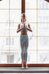 Slim beautiful young girl in sportswear is standing at the window and meditating with closed eyes. Concept of relaxation and spirituality. Yoga concept