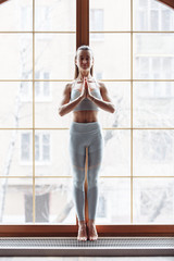 Fototapeta premium Slim beautiful young girl in sportswear is standing at the window and meditating with closed eyes. Concept of relaxation and spirituality. Yoga concept