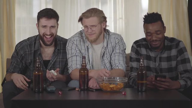 Three happy men friends sitting at house on the cauch, looking tv, drinking beer with chips, having a video call to a someone.