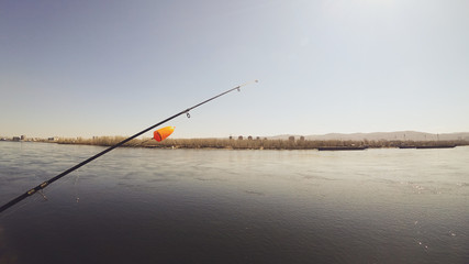Fishing rod with a bright orange large float. Background river, ships, houses and mountains on the opposite shore. Bright sun.  