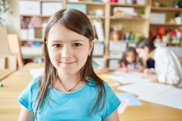 Sweet girl smiling and looking at camera while standing on blurred background of classroom in art school