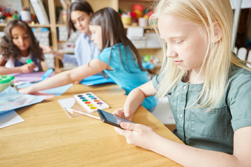 Albino girl using modern smartphone while sitting at desk during lesson in art school, teacher and...