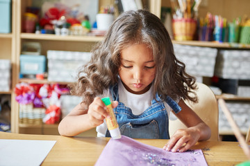 Sweet girl with curly hair applying glue stick on paper sheet with glitter during lesson in art...