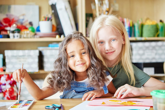 Two sweet girls smiling and looking at camera while painting nice picture during lesson in art school