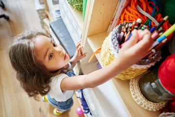 From above shot of sweet little girl taking colored pencils from shelf in art school classroom