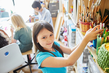 Side view of cute girl smiling and looking at camera while standing near shelf with various drawing...