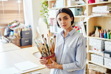 Portrait of young teacher holding basket with set of brushes and looking at camera while standing in art classroom in school