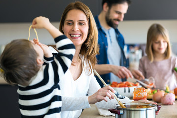 Cute little boy having fun with his young mother while eating spaghetti on kitchen at home.