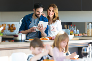 Attractive young couple using digital tablet while having lunch with their sons in the kitchen at home.