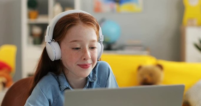Close up of the cute and pretty red haired small schoolgirl in the headphones smiling and watching something on the laptop computer at home.