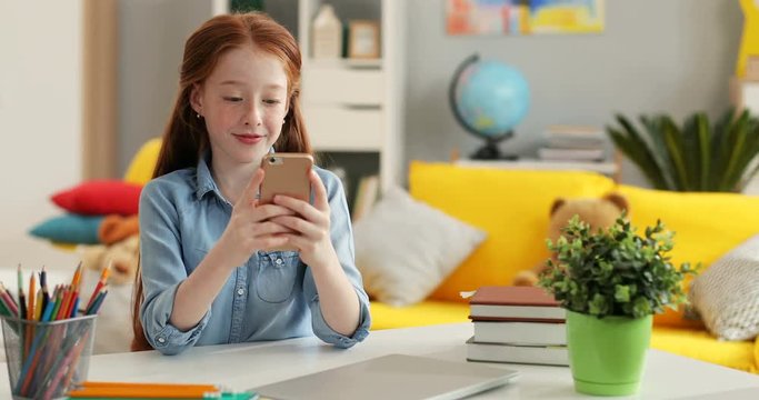 Nice Caucasian schoolgirl with long red hair sitting at the desk in her cozy room and tapping or scrolling on the smartphone.