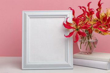 Composition from a decorative wooden frame and red and yellow flowers of Glorios in a glass vase on a white shelf on a pink background