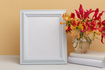 Composition from a decorative wooden frame and red and yellow flowers of Glorios in a glass vase on a white shelf on a yellow background