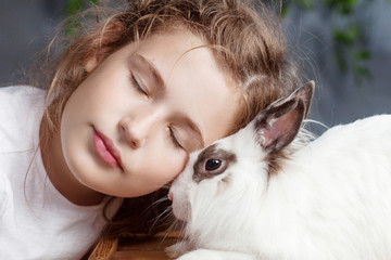 Little girl playing with real rabbit. Child and white bunny on Easter on flower background. Kid kiss pet. Fun and friendship for animals and children.