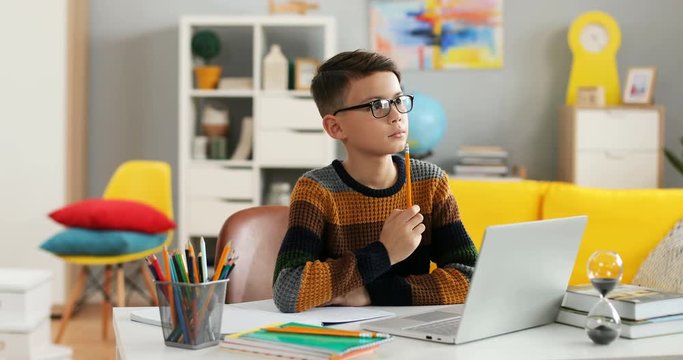 Portrait shot of the Caucasian small cute teen boy writing exercise of homework in the copybook and thinking while sitting at the table with a laptop computer at home.