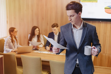A stylish man in a jacket and a shirt with a cup of coffee in his hand stands and reads documents on the background of working colleagues at office