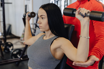 Fototapeta premium A young girl wearing sportswear in a gym performs dumbbell exercises, the coach helps her