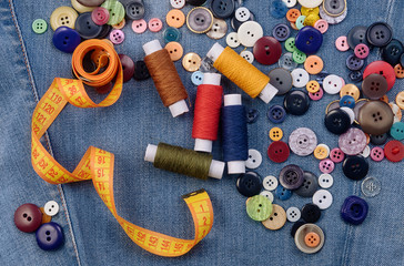 Fototapeta na wymiar A pile of different sewing buttons, yellow measuring tape and thread spools on denim jeans background. Flat lay.