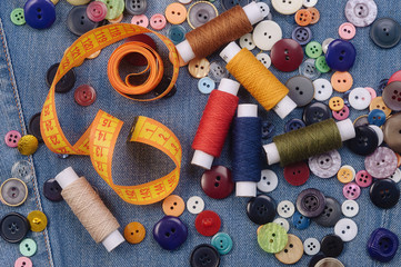 Fototapeta na wymiar A pile of multicolored sewing buttons, yellow measuring tape and thread spools on denim jeans background. Flat lay.