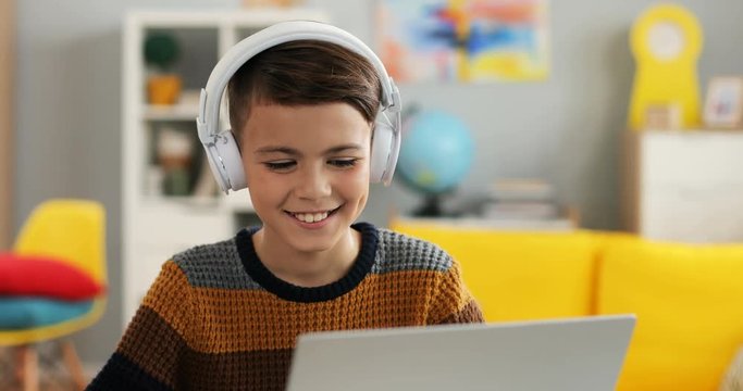 Close up of the Caucasian little cute teen boy in headphones tapping and playing on the laptop computer in his room.