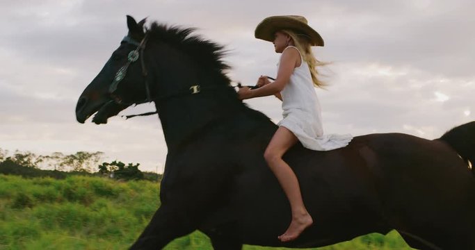 Majestic girl in white dress horseback riding with galloping black horse in slow motion at sunset
