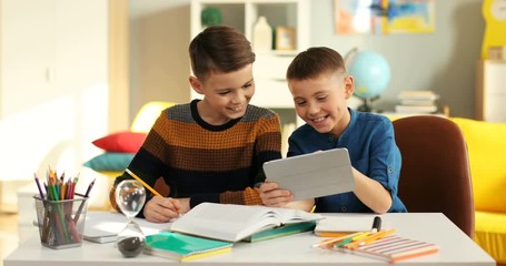 Little Caucasian cute teen boy showing to his best friend something on the tablet computer in the kid's room at the table.