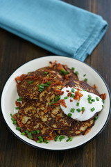 Potato pancakes draniki served with sour cream, chives and bacon on a white plate. Dark wooden table, blue linen napkin, high resolution