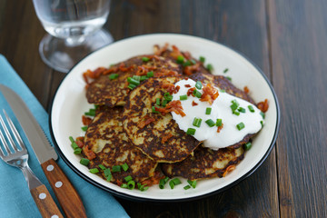 Potato pancakes draniki served with sour cream, chives and bacon on a white plate. Dark wooden table, blue linen napkin, high resolution