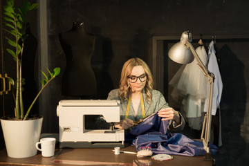 Obraz na płótnie Canvas beautiful young seamstress girl in glasses in a jacket at work sews a dress on a sewing machine in a dark room at night