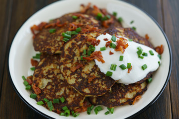 Potato pancakes draniki served with sour cream, chives and bacon on a white plate. Dark wooden table, blue linen napkin, high resolution