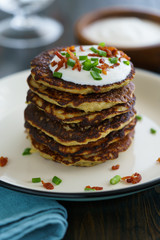 Potato pancakes draniki served with sour cream, chives and bacon on a white plate. Dark wooden table, blue linen napkin, high resolution