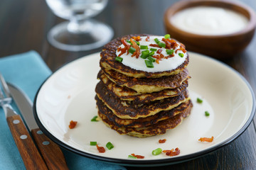Potato pancakes draniki served with sour cream, chives and bacon on a white plate. Dark wooden table, blue linen napkin, high resolution