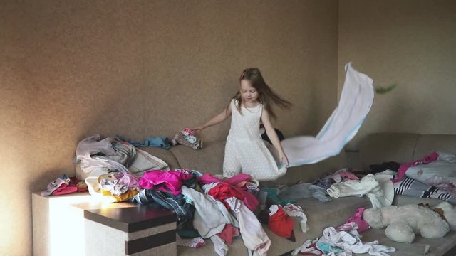 Funny playful girl in dress having fun throwing clothes or making mess on sofa at home while parents are away.