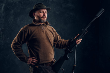 Confident hunter posing with a rifle and looking sideways. Studio photo against dark wall background