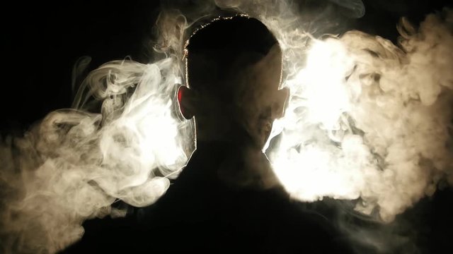 Young guy vaping in the dark room. Silhouette of vaper in the darkness lighted by bright lamps. 